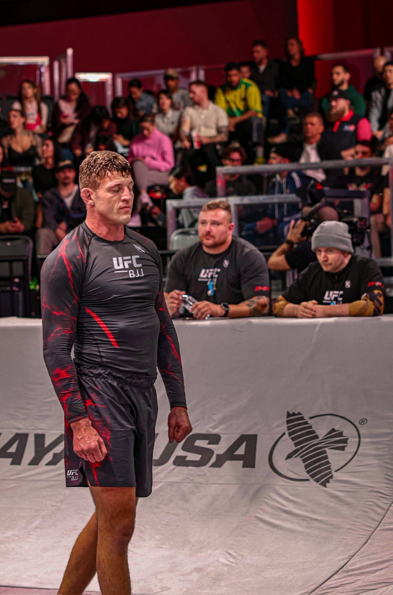 Nicholas Meregali in UFC BJJ red rashguard standing at ringside, packed arena crowd behind him