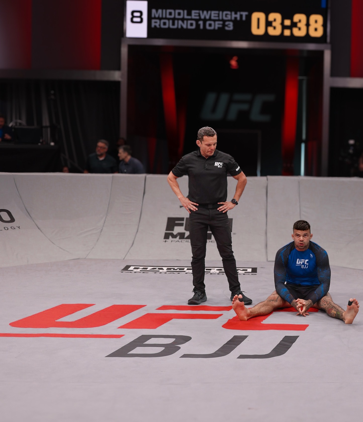 UFC BJJ athlete sitting on the mat after a tough middleweight bout, referee standing over him